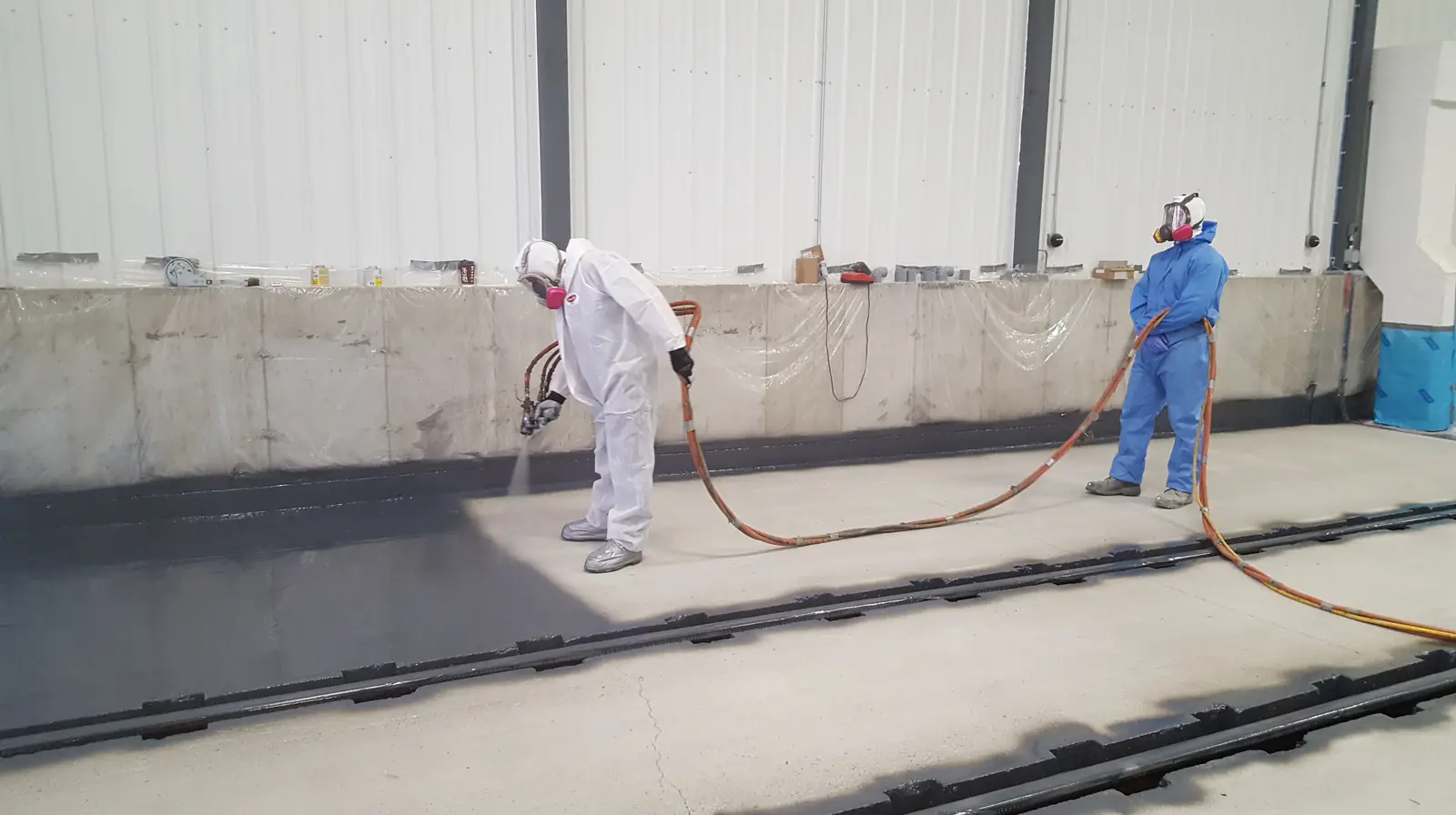 Two applicators spraying a long concrete wall with hoses running across the floor